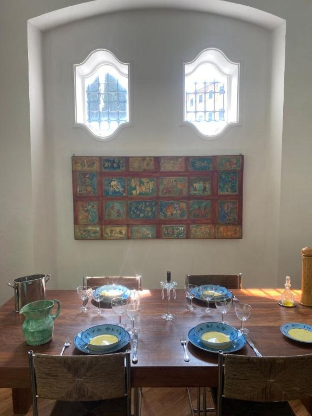 A dining area with a wooden table set for four, two windows above, and a wall with colorful artwork.