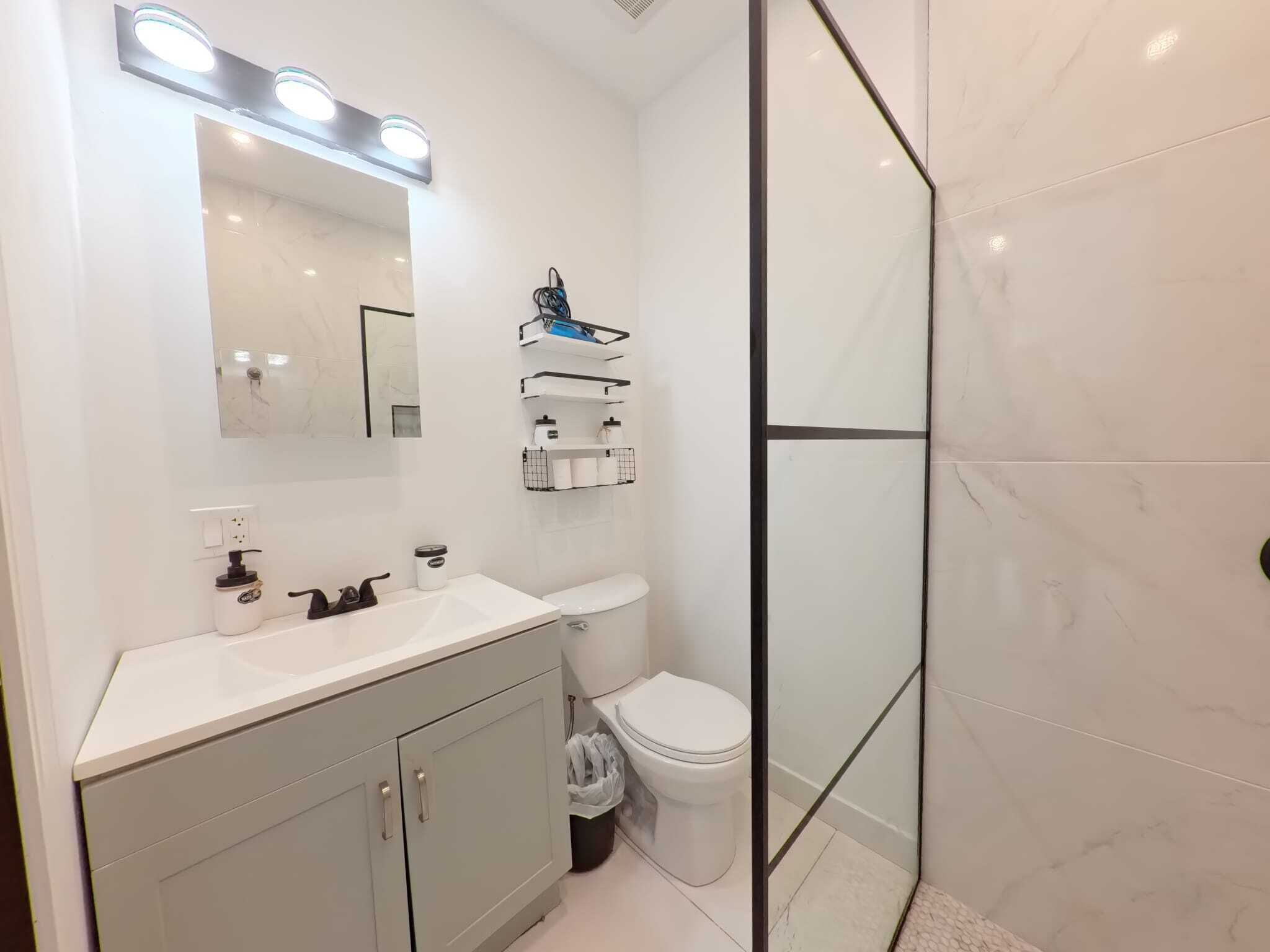 A modern bathroom with a white vanity, a toilet, a glass shower enclosure, and a mirror with lights.