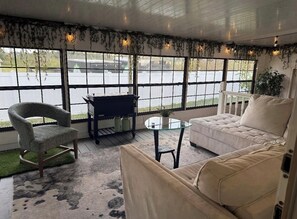 A cozy room with a sofa, armchair, and a small table, large windows with a view of the parking lot, and a ceiling with hanging plants.