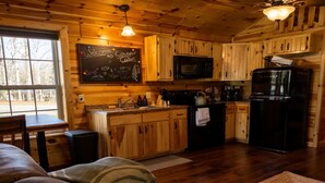 Une cuisine chaleureuse, aménagée dans le style d’un chalet, avec des meubles en bois, un réfrigérateur noir et un tableau noir sur lequel est écrit un message de bienvenue.