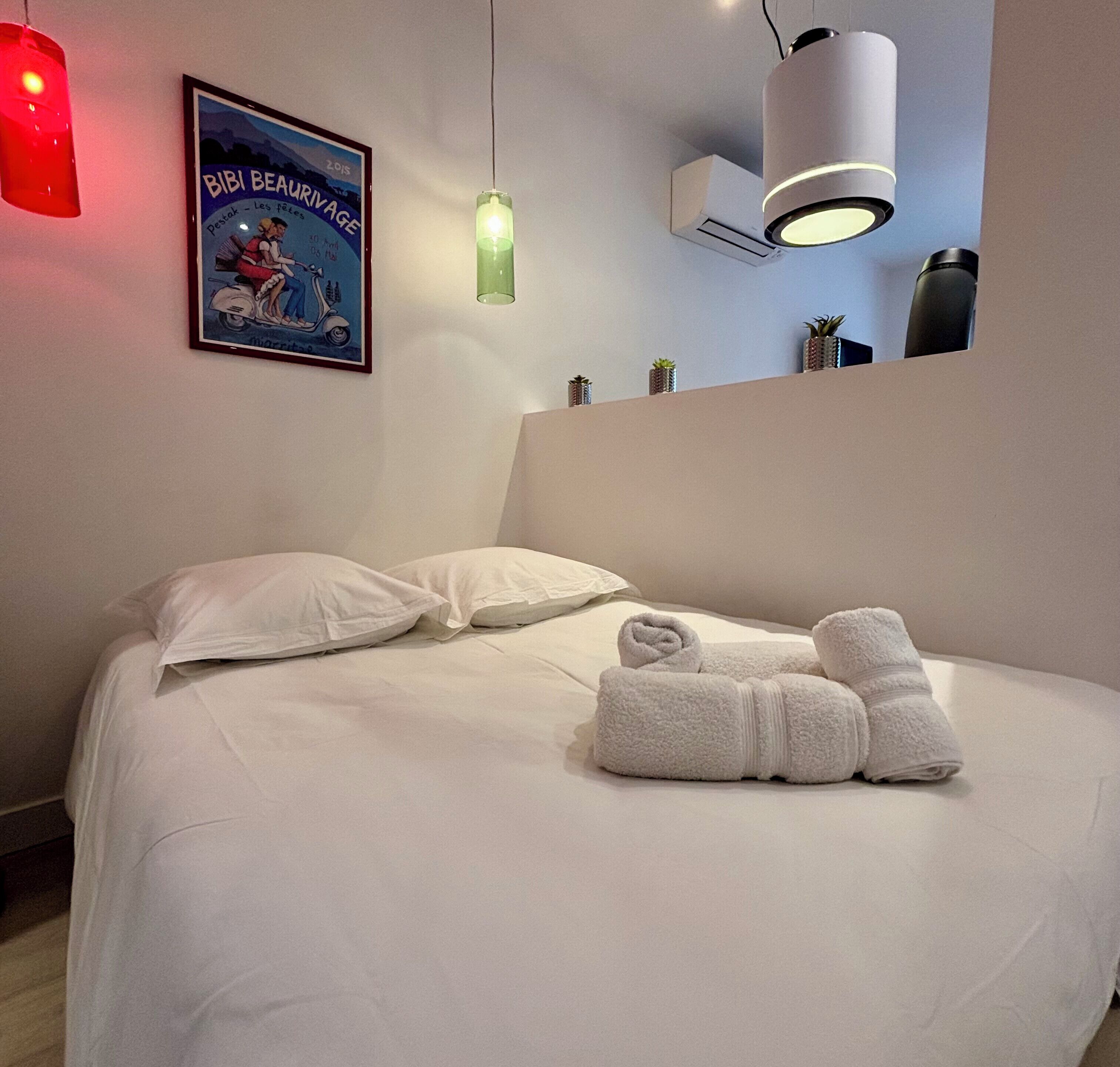 Una cama bien hecha con almohadas y toallas enrolladas, un cartel enmarcado en la pared y una lámpara colgante.