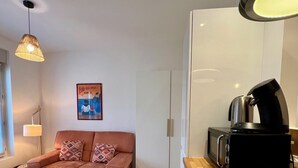 A compact living space with a kitchenette, a sofa with patterned cushions, a hanging light fixture, and a framed picture on the wall. - CASA BLUM #2 - Studio climatisé, à 2 pas de la gare (Brive-la-Gaillarde)