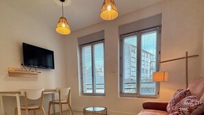 A modern living room with a sofa, a TV mounted on the wall, and two windows with blinds. - CASA BLUM #2 - Studio climatisé, à 2 pas de la gare (Brive-la-Gaillarde)