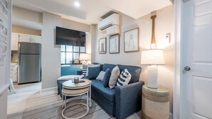 A modern living room with a sofa, a small round table, and a lamp. There is a kitchen area in the background with a refrigerator and a window. - Beach Access, Hot Tub, Ping Pong Table, Bikes! (Tybee Island)