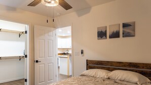 Een slaapkamer met een bed, een houten hoofdbord en een plafondventilator. Er zijn drie aan de muur bevestigde foto’s en een deur die leidt naar een keukenhoek.