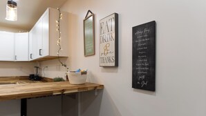 Een keuken met witte kastjes, een houten aanrechtblad en een mand met spullen. Er hangen drie decoraties aan de muur: één met 'EAT DRINK & THANKS', een andere met 'Always Ping', en een derde met een hart en een citaat.