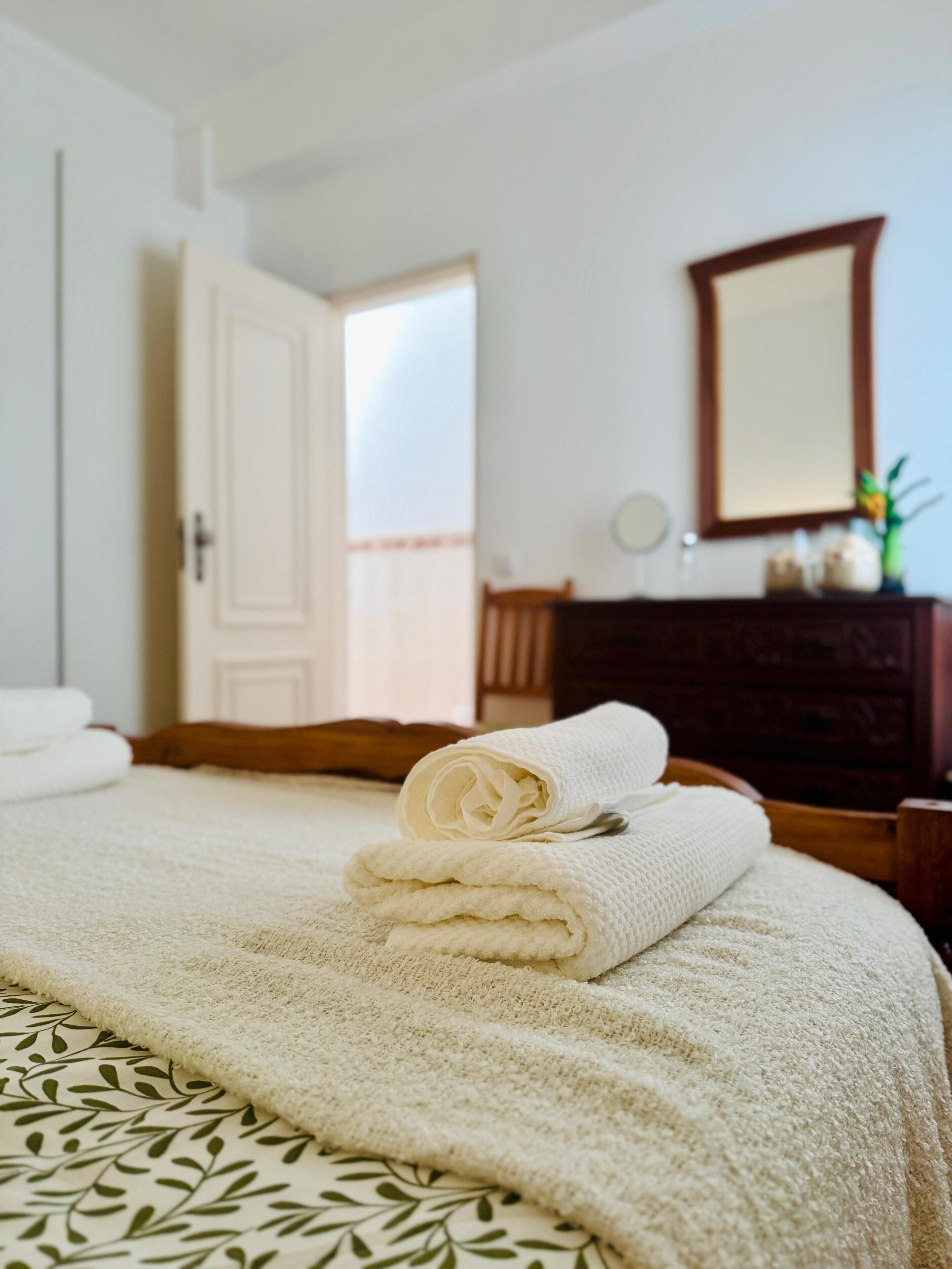 A neatly made bed with a rolled white towel, a wooden dresser with a mirror, and a door leading to an outdoor area.