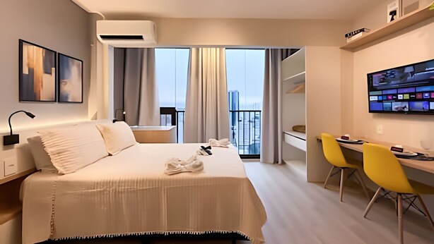 Une chambre d’hôtel moderne dotée d’un grand lit, d’un bureau avec des chaises jaunes, d’un téléviseur à écran plat et d’un balcon avec des rideaux.