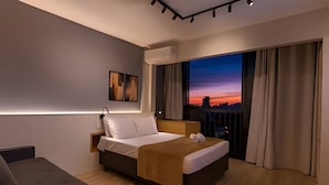 Une chambre d’hôtel moderne, équipée d’un lit, d’une table de chevet, d’une lampe, offrant une vue sur la ville au coucher du soleil, et dotée d’une fenêtre avec des rideaux.