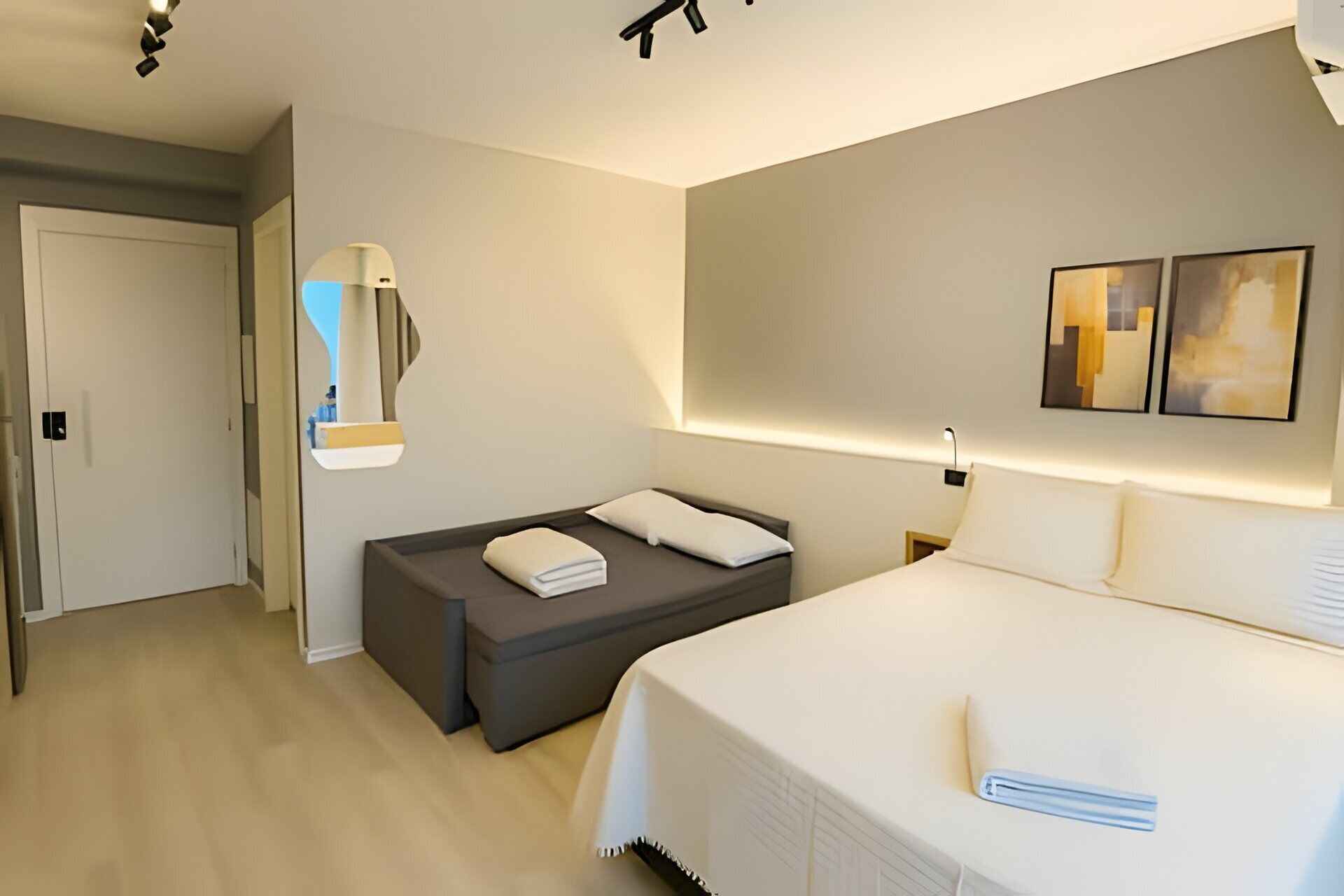A modern hotel room with a large bed, a smaller sofa, a bedside table, a mirror, and two wall-mounted artworks.