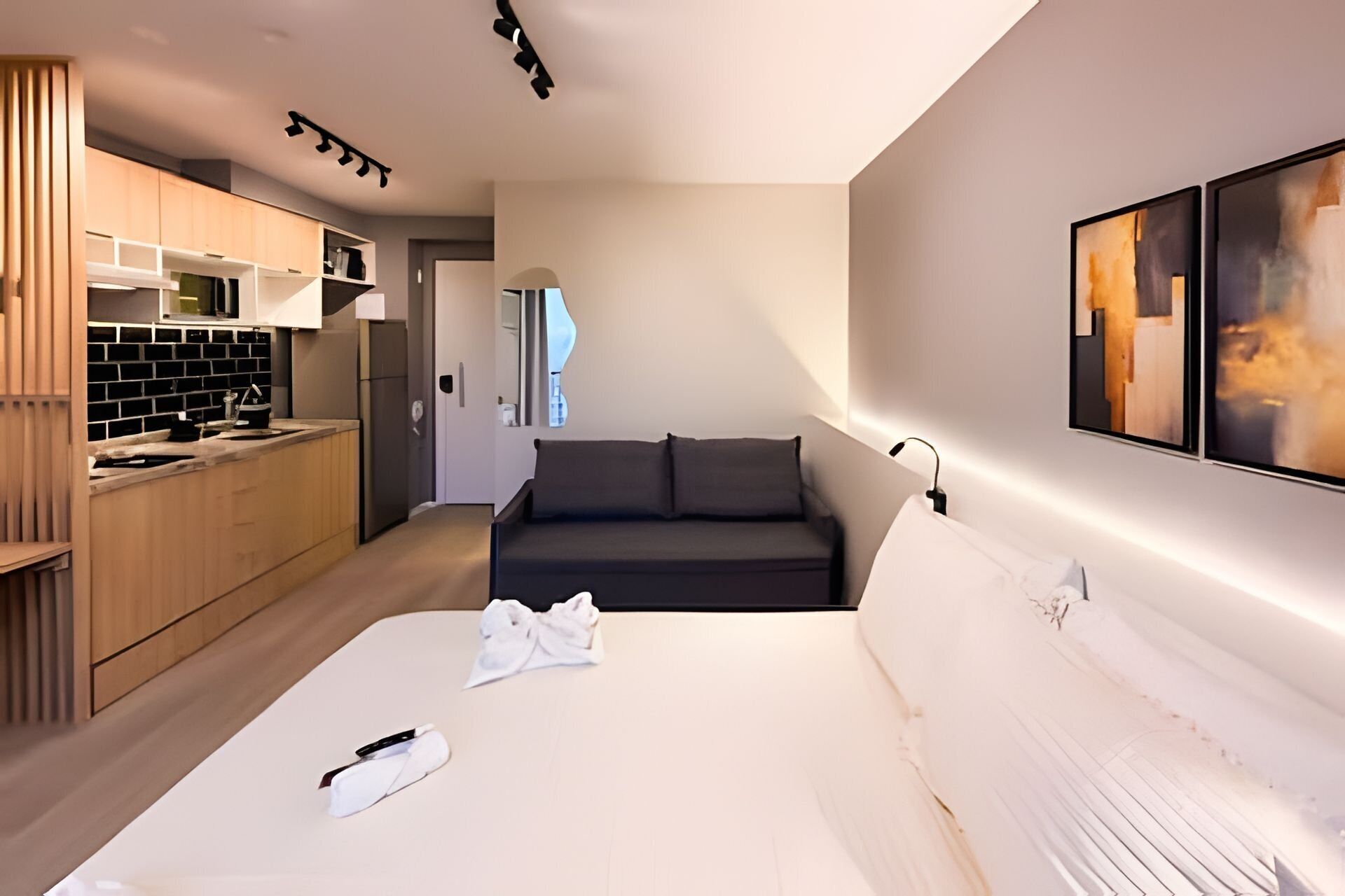 A modern hotel room with a bed, a sofa, a kitchen area, and artwork on the wall.