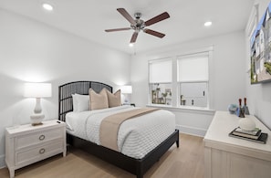 Un dormitorio con cama, mesitas de noche, escritorio, ventilador de techo y ventana con vista a edificios.