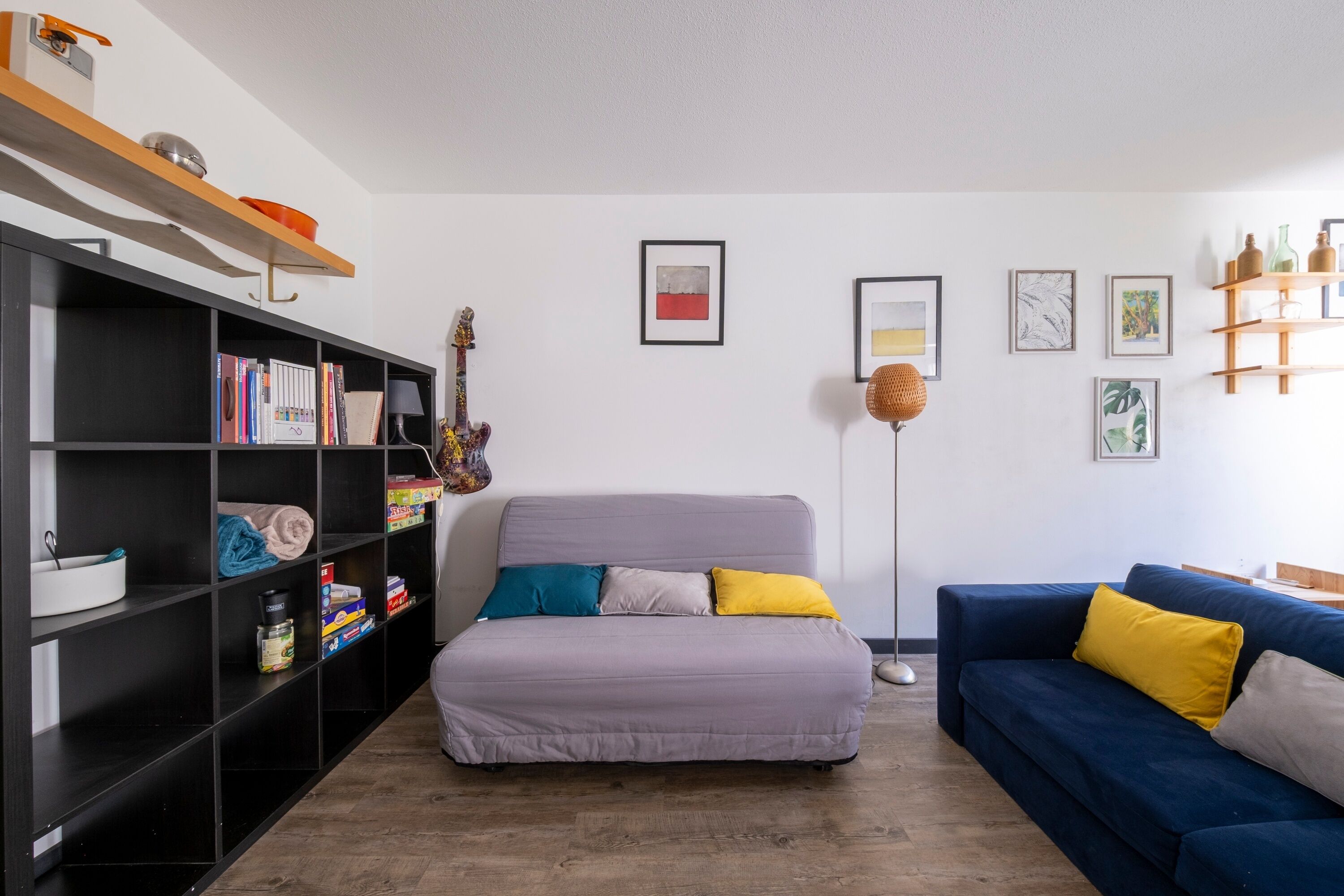 A modern living room with a grey sofa, a blue sofa with yellow cushions, a bookshelf, and a floor lamp.