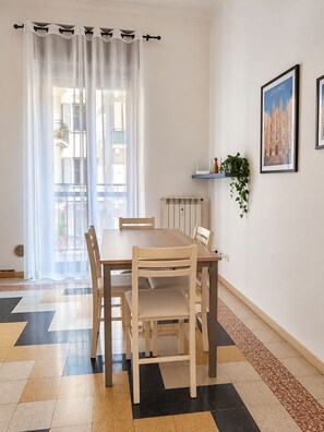 Een eethoek met een tafel en stoelen, een radiator, een raam met gordijnen en een lijst met een afbeelding aan de muur.