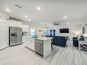 A modern kitchen with white cabinetry, a stainless steel refrigerator, and a dishwasher. There is a blue sofa and a television in the living area. - Salty Joe's-WindMark Beach-Pools-Golf Cart-Sleeps 10-InLaw Suite (Port St. Joe)