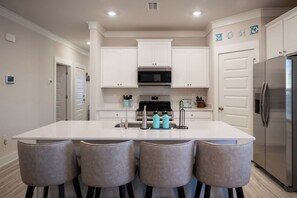 A modern kitchen with white cabinets, a stainless steel refrigerator, a microwave, and a large island with bar stools. - Salty Joe's-WindMark Beach-Pools-Golf Cart-Sleeps 10-InLaw Suite (Port St. Joe)