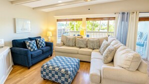 A spacious living room with a blue sofa, a beige sofa, and a matching ottoman. There is a window with a view of greenery outside. - Unique 2 in 1  * BayViews * Pool (South Padre Island)