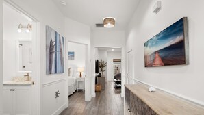 A hallway with a wooden floor, white walls, and a wooden bench. There are two paintings on the walls, a sink with a marble countertop, and a door leading to another room. - Beach House - Sunny Sands  (Panama City Beach)