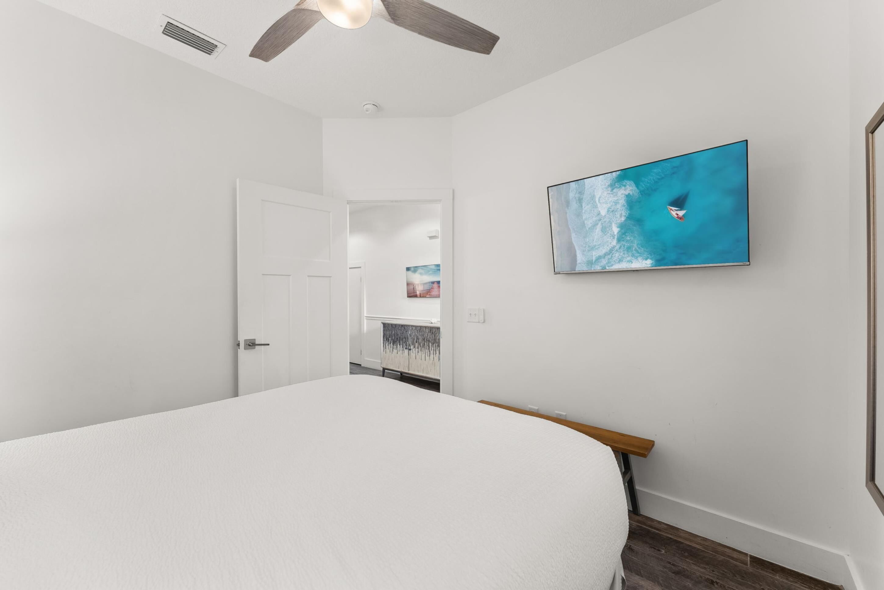 A bedroom with a bed, a ceiling fan, a wall-mounted TV, and a door leading to another room.