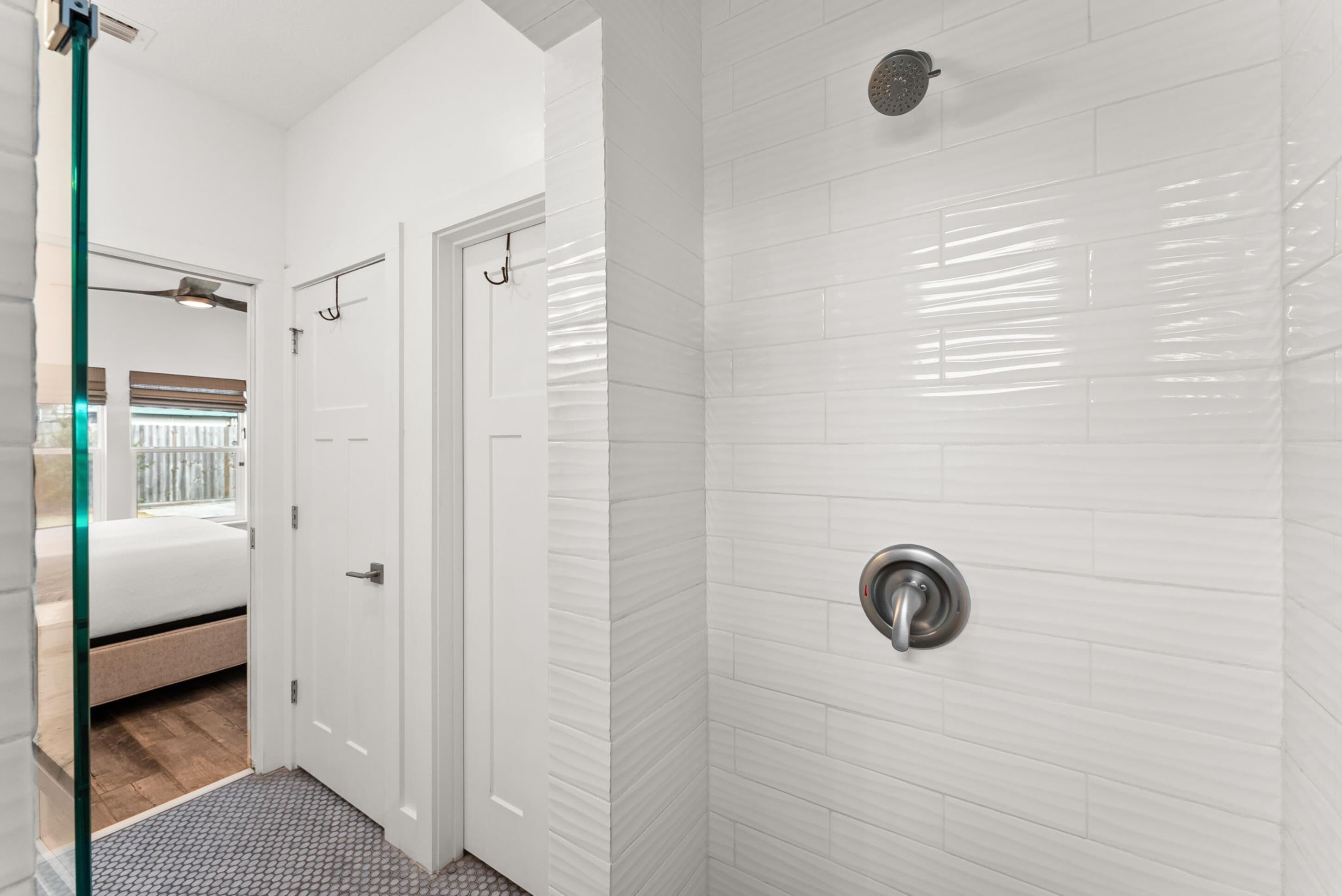 Un bagno moderno con pareti piastrellate di bianco, una doccia con soffione manuale e una porta in vetro che dà su una camera da letto con un letto e una finestra.