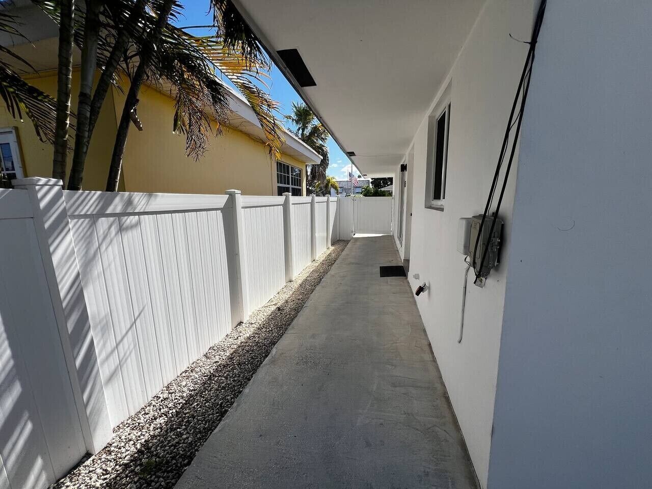 A narrow concrete pathway flanked by white fences, leading to a building with a flat roof and a small window.