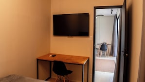 Ett sovrum med en säng, ett skrivbord och en stol. Det finns en tv som är monterad på väggen och en dörr som leder till ett annat rum.