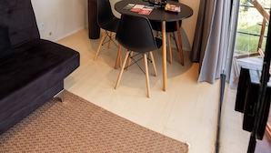 Ett modernt vardagsrum med en svart soffa, ett matbord med stolar, en krukväxt och en utsikt genom ett stort fönster.