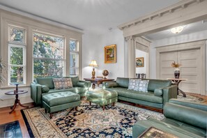 Ein geräumiges Wohnzimmer mit einem grünen Ecksofa, einem gemusterten Teppich und einem großen Fenster mit Jalousien.