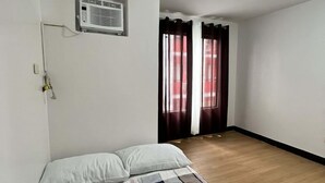 Une chambre à coucher avec un lit, une fenêtre avec des rideaux, un climatiseur fixé au mur et un plancher en bois.
