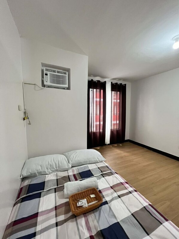A bedroom with a bed, a window with curtains, a wall-mounted air conditioner, and a wooden floor.
