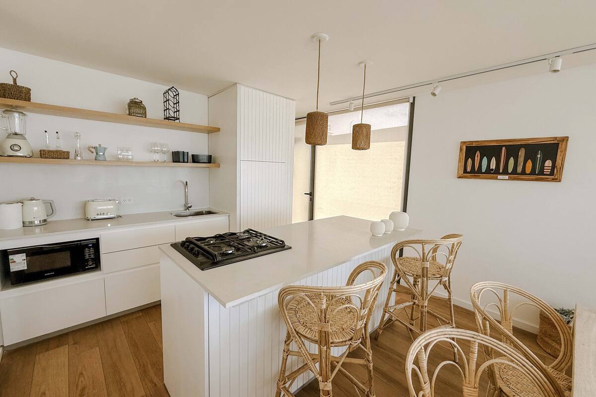 A modern kitchen with white cabinetry, a central island, wicker chairs, and a dining table.