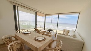 A dining area with a wooden table and wicker chairs, a sofa, and large windows overlooking the beach. - Punta Cantera Suites de Mar (Mar del Plata)