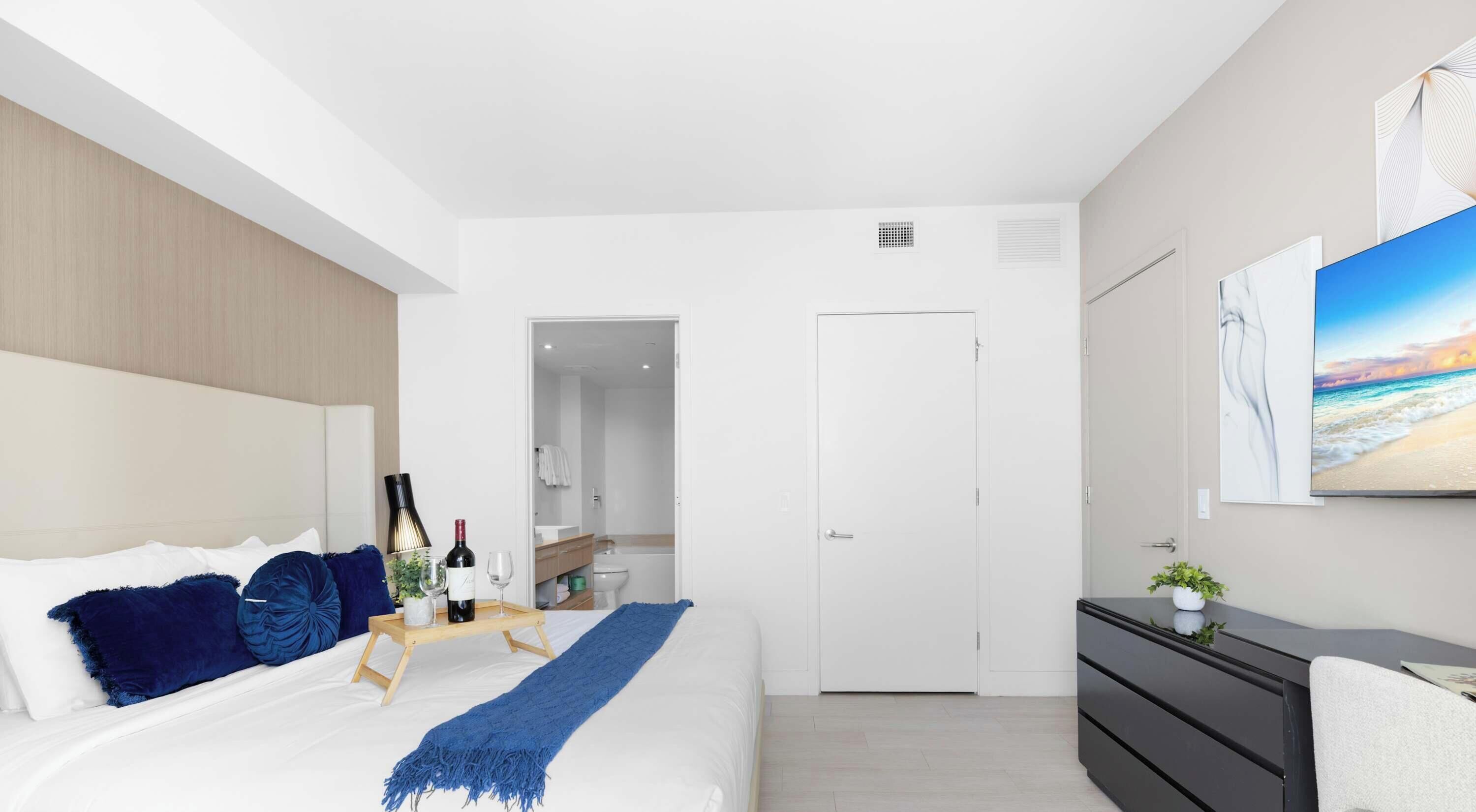 A modern bedroom with a bed, bedside table, dresser, and a painting of a beach scene.