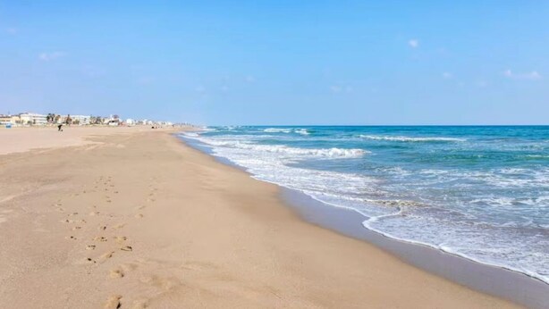 A wide sandy beach with gentle waves, a clear sky, and buildings in the distance.