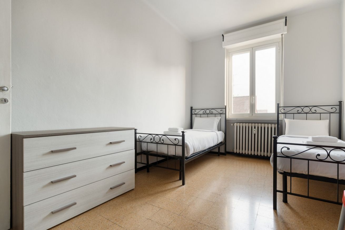 A bedroom with two single beds, a dresser, a radiator, and a window with blinds.