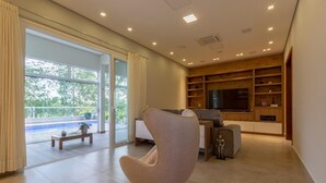 A modern living room with a large window, a sofa, a TV, and built-in shelves. - Mansion in Greater São Paulo Area - Condominium (Caconde)