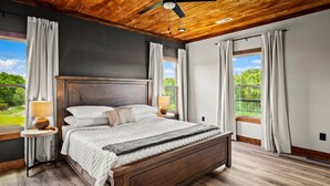A bedroom with a wooden ceiling, a large bed with a headboard, two bedside tables with lamps, and a view of greenery through large windows. - New 2025–5 Mi Dollywood–Mtn Views–1-Step–Pets (Sevierville)