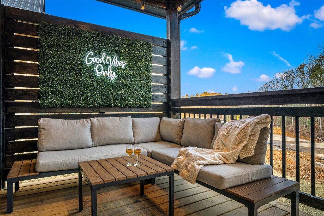 A cozy outdoor seating area with a wooden bench, a cushioned sofa, and a table with two glasses of wine. A neon sign reads "Good Vibes Only."