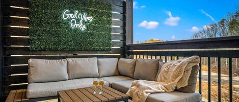 A cozy outdoor seating area with a wooden bench, a cushioned sofa, and a table with two glasses of wine. A neon sign reads "Good Vibes Only."