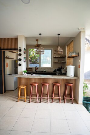 A modern kitchen with a central island, bar stools, pendant lights, and a view of the outdoors.