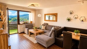 Un salon moderne avec un canapé, une table basse et un coin repas, de grandes fenêtres donnant sur un espace verdoyant et un miroir au mur.