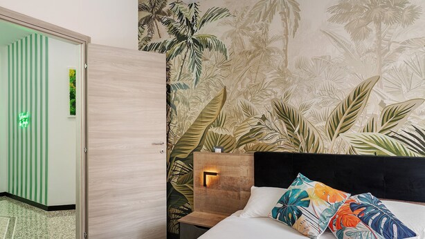 Ein Hotelzimmer mit Bett, Nachttisch, einer Wand mit tropischem Wandpapier, einer Tür und einem kleinen Tisch.