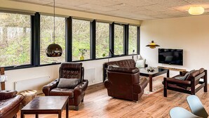 Een moderne woonkamer met lederen banken, een houten salontafel en een televisie. Grote ramen bieden uitzicht op bomen.