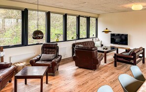 A modern living room with leather sofas, a wooden coffee table, and a TV. Large windows offer a view of trees. - 3 bedroom stunning home in Struer (Struer)