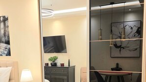 A modern living room with a large mirror, a flat-screen TV, a sofa, a small side table, and a dining area with chairs and a table. - Elegant Apartment in Belgrade City Center – Stari Grad (Beograd)