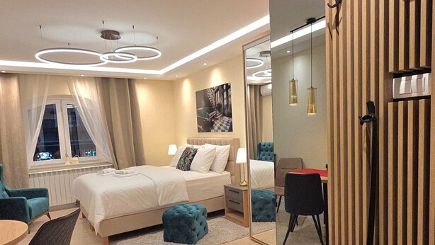 A modern hotel room with a large bed, a sitting area with a table and chairs, a mirror, and a view of the outside. - Elegant Apartment in Belgrade City Center – Stari Grad (Beograd)