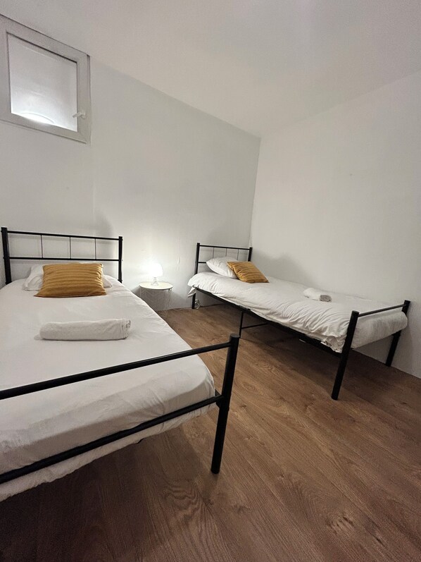 A room with two single beds, a small bedside table with a lamp, and a window with a view of the sky.