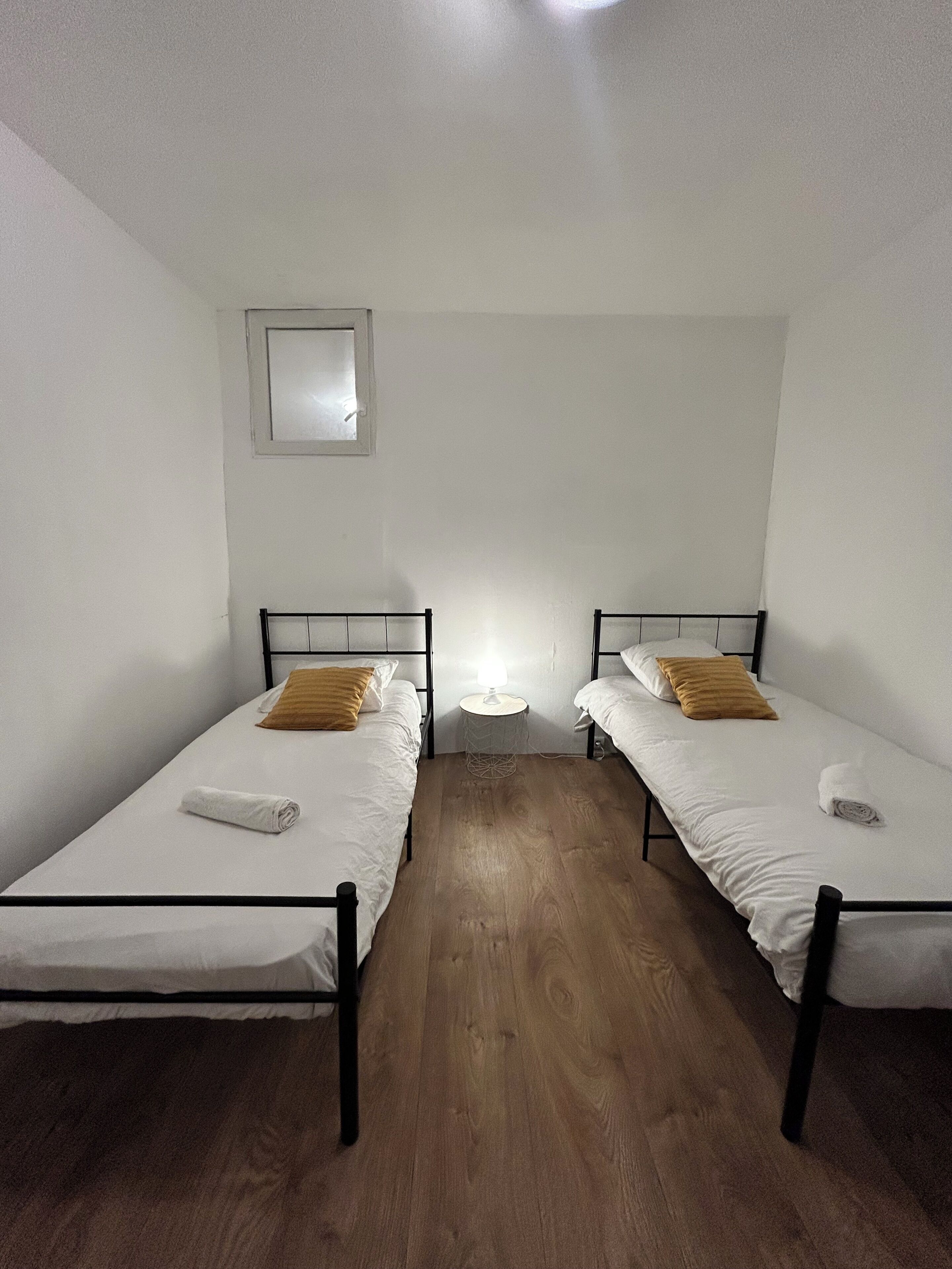 A small, clean room with two single beds, a small window, and a bedside table with a lamp.