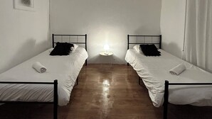 Une petite pièce à la lumière tamisée, avec deux lits simples, un plafonnier rond et une petite table avec une lampe.