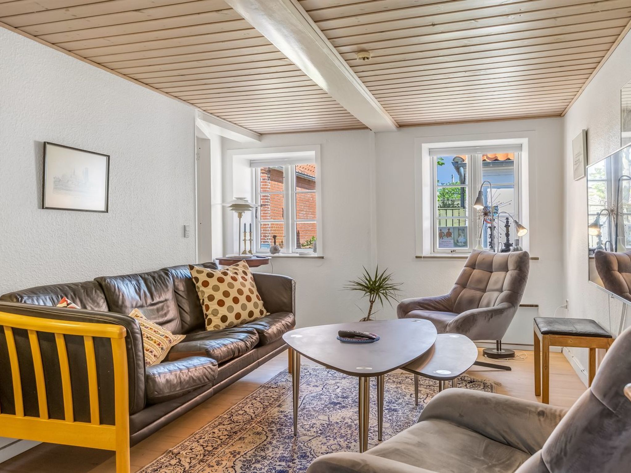 Una sala moderna con un sofá de cuero, un sillón a juego, una pequeña mesa de centro y un banco de madera. Hay dos ventanas que proporcionan luz natural y vistas al exterior.